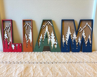 May include: Wooden letters spelling "LIAM" with a mountain and forest design. The letters are red, orange, green, and blue, with white mountain and tree cutouts. The letters are placed on a cream-colored knitted surface.
