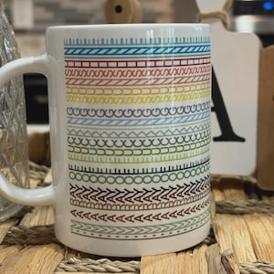 May include: White ceramic mug with a colorful geometric pattern. The design features horizontal bands of various shapes and colors, including blue, red, yellow, and green. The mug has a curved handle and is set against a neutral background.