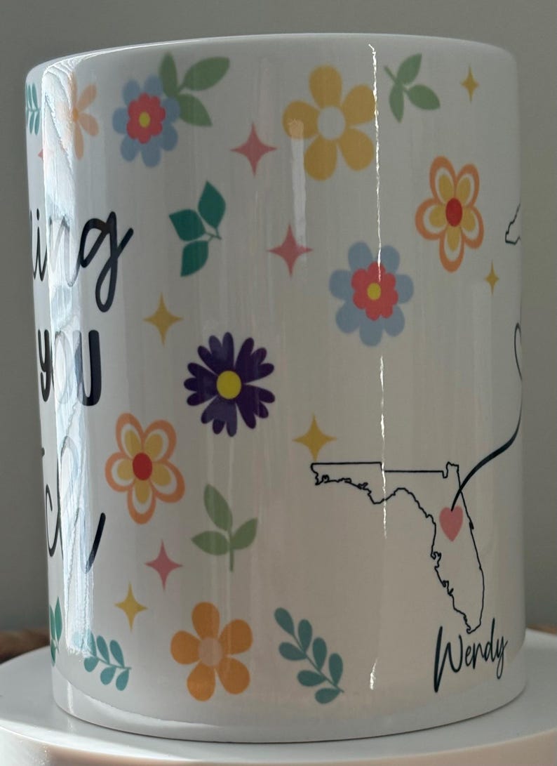 Pode incluir: Caneca de cer&acirc;mica branca decorada com flores coloridas, estrelas e folhas. A caneca apresenta o texto "Missing You" e um contorno do estado da Fl&oacute;rida com um cora&ccedil;&atilde;o. O nome "Wendy" tamb&eacute;m &eacute; exibido.