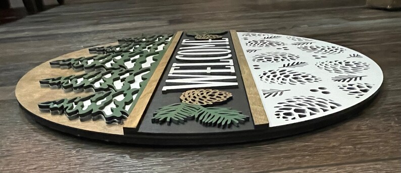 May include: A circular wooden welcome sign with a nature theme. The sign features a tree design, the word "WELCOME", and a pine cone design. The sign is painted in green, white, brown, and black. The diameter is approximately 30 cm.
