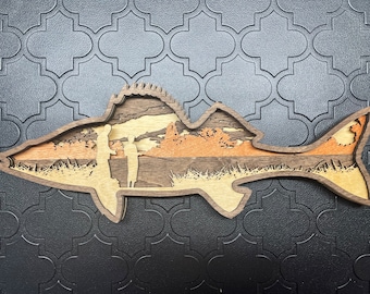 May include: A layered wooden fish-shaped wall art piece. The artwork features a landscape scene with two figures, trees, and a sunset. The fish is made of various shades of brown and tan wood.