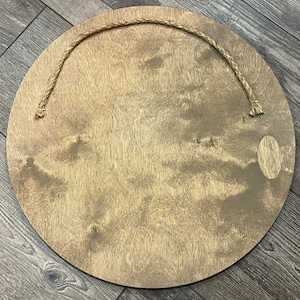May include: A circular wooden sign with a natural wood grain finish. A jute rope is attached at the top for hanging. The sign has a light brown colour and a smooth surface.