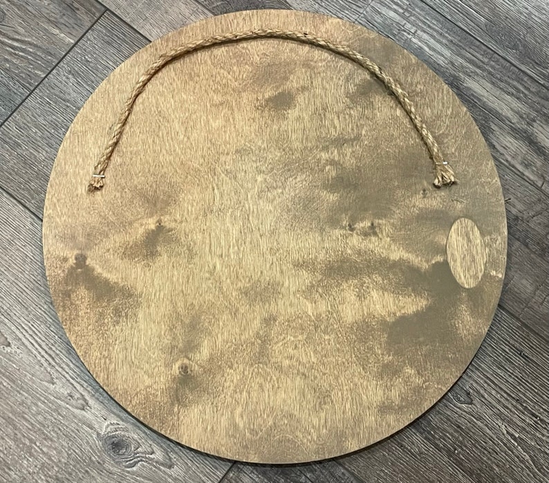 May include: A circular wooden sign with a natural wood grain finish. A jute rope is attached at the top for hanging. The sign has a light brown colour and a smooth surface.