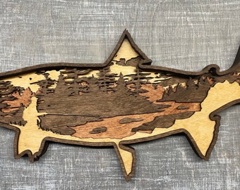 May include: A wooden fish-shaped wall art piece. The layered design features a landscape scene with trees, a river, and mountains. The artwork uses natural wood tones of light tan, brown, and reddish-brown.