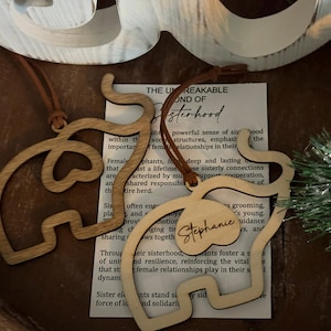 Op de afbeelding: Twee houten olifantvormige ornamenten met leren koorden hangen in de buurt van een bedrukt papier. E&eacute;n ornament heeft de naam "Stephanie" gegraveerd. Het papier bevat tekst over zusterschap. De ornamenten zijn lichtbruin.