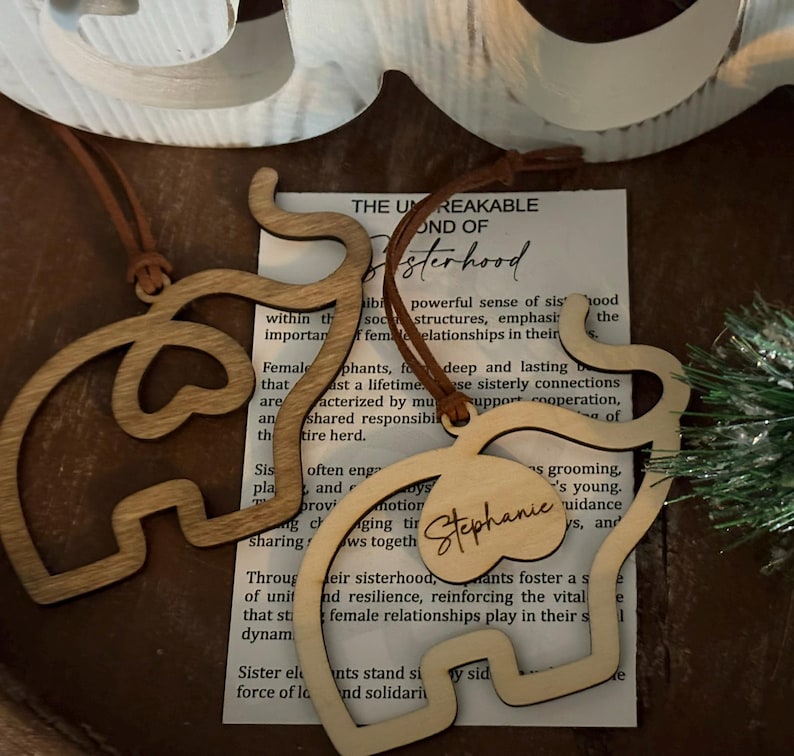 Op de afbeelding: Twee houten olifantvormige ornamenten met leren koorden hangen in de buurt van een bedrukt papier. E&eacute;n ornament heeft de naam "Stephanie" gegraveerd. Het papier bevat tekst over zusterschap. De ornamenten zijn lichtbruin.