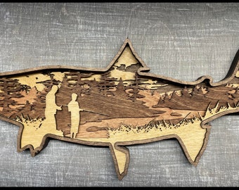 May include: A layered wooden art piece in the shape of a fish. The interior depicts a fishing scene with two figures, trees, and a river. The wood has natural tones of light and dark brown.