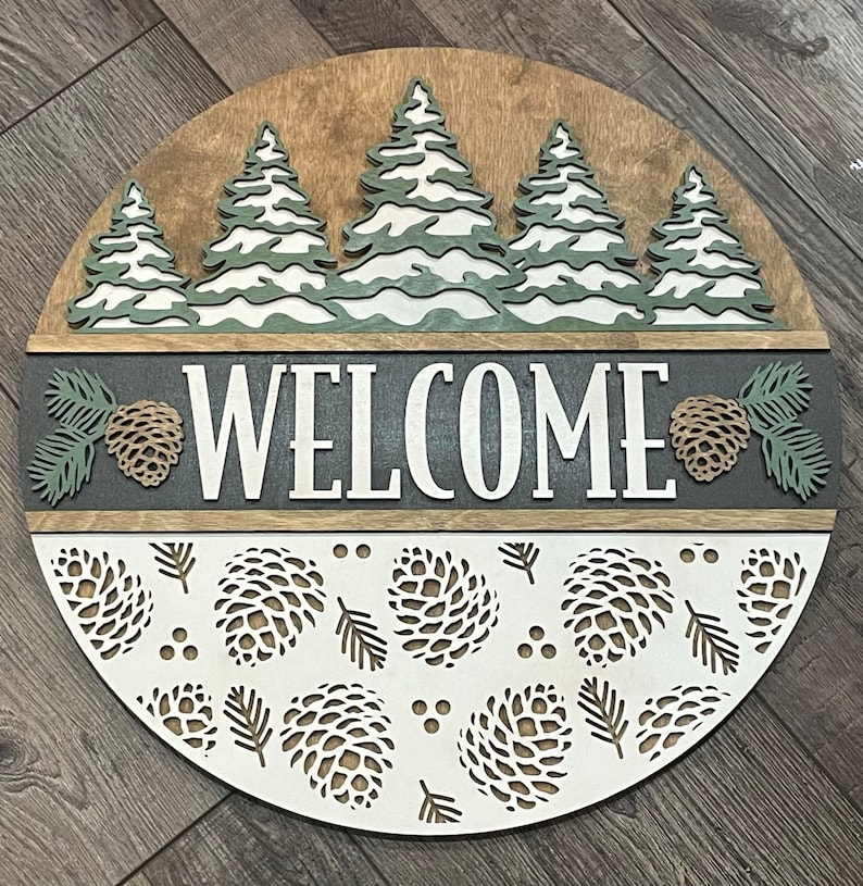 May include: A circular wooden welcome sign with a forest theme. The sign features layered pine trees, the word "WELCOME", pinecones, and leaf cutouts. The colour palette includes shades of brown, white, grey, and green.