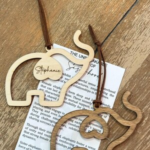 Op de afbeelding: Twee houten olifantvormige ornamenten met bruine leren koorden. E&eacute;n is lichtgekleurd met de naam "Stephanie" en de andere is donkerder hout. Een papier met tekst is zichtbaar.