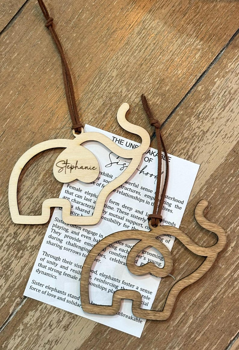 Op de afbeelding: Twee houten olifantvormige ornamenten met bruine leren koorden. E&eacute;n is lichtgekleurd met de naam "Stephanie" en de andere is donkerder hout. Een papier met tekst is zichtbaar.