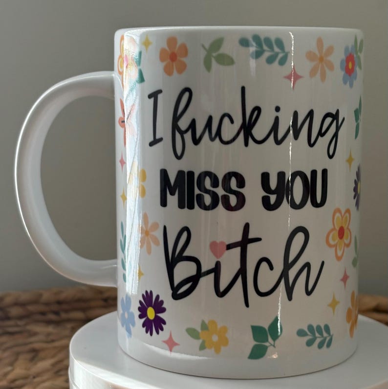 Pode incluir: Caneca de cer&acirc;mica branca com o texto "I fucking MISS YOU Bitch" em escrita preta. A caneca &eacute; decorada com desenhos florais e de estrelas coloridas. A caneca tem uma al&ccedil;a branca.