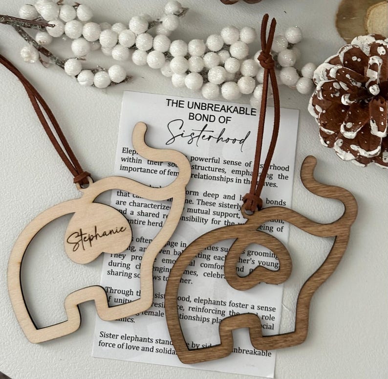 Op de afbeelding: Twee houten olifantvormige ornamenten met leren koorden om op te hangen. E&eacute;n is lichtgekleurd met de naam "Stephanie" en de andere is donkerder hout. De ornamenten worden getoond met een kaart met de tekst "The Unbreakable Bond of Sisterhood."