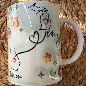 Pode incluir: Caneca de cer&acirc;mica branca decorada com desenhos florais coloridos e contornos de estados. A caneca apresenta um cora&ccedil;&atilde;o que conecta os contornos dos estados de Michigan e Ge&oacute;rgia, com os nomes "Brittany" e "Melissa" escritos abaixo.