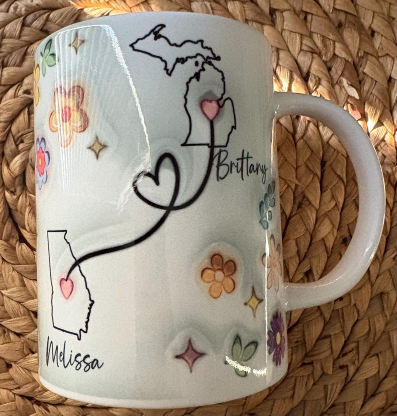Pode incluir: Caneca de cer&acirc;mica branca decorada com desenhos florais coloridos e contornos de estados. A caneca apresenta um cora&ccedil;&atilde;o que conecta os contornos dos estados de Michigan e Ge&oacute;rgia, com os nomes "Brittany" e "Melissa" escritos abaixo.