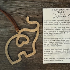 Op de afbeelding: Houten olifantenornament met een bruin leren bandje, naast een kaart met de tekst "THE UNBREAKABLE BOND OF Sisterhood". Het ornament is lichtbruin en heeft een uitgesneden ontwerp. De kaart beschrijft vrouwelijke relaties.