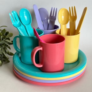 May include: A set of colorful plastic cups and utensils, including forks, spoons, and a knife, arranged on a stack of colorful plates. The cups and utensils are all different colors, including blue, yellow, pink, and purple.