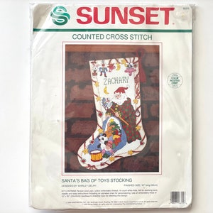 May include: A counted cross stitch pattern for a Christmas stocking featuring Santa Claus with a bag of toys. The stocking is white with red and green trim and the name "Zachary" is embroidered on the front. The pattern includes a color-coded chart and instructions for stitching.