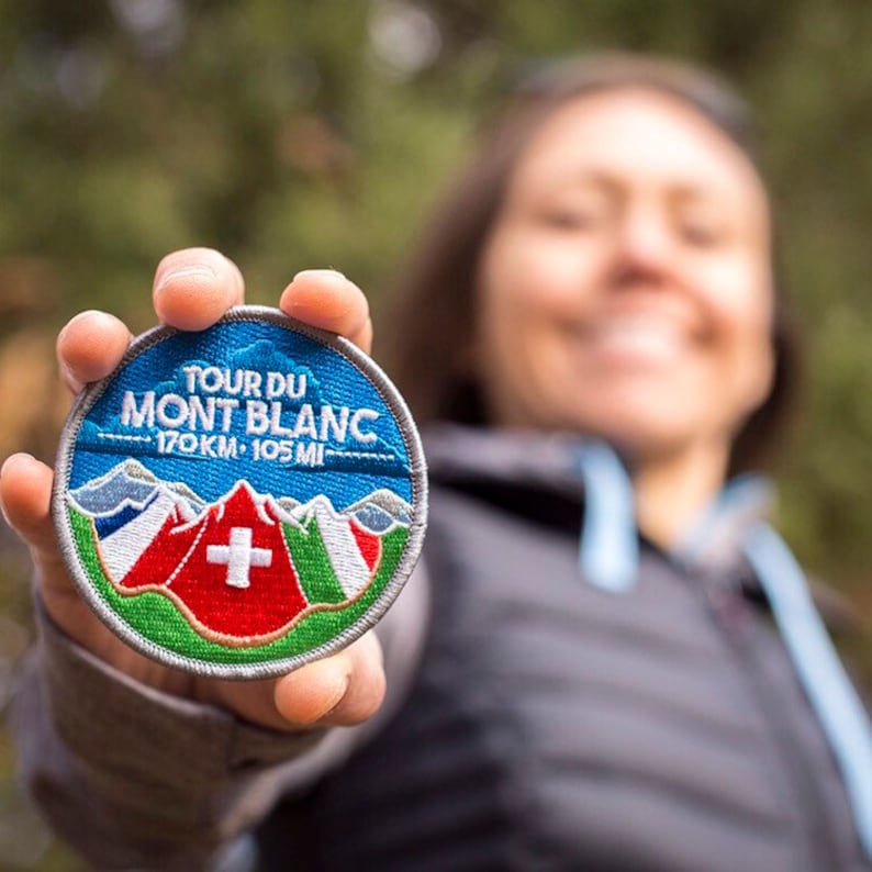 May include: A circular embroidered patch with the text "TOUR DU MONT BLANC" and the distance "170KM - 105 MI". The patch features a mountain range, the Swiss flag, and a blue border.