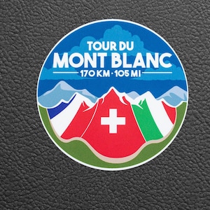 Könnte beinhalten: Runder Aufkleber mit einem Bergdesign unter blauem Himmel. Der Text "TOUR DU MONT BLANC" steht über den Bergen, darunter "170 KM • 105 MI". Der Aufkleber zeigt die Flaggen von Frankreich, der Schweiz und Italien.