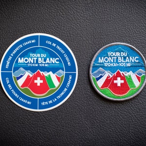 Könnte beinhalten: Zwei runde blaue und weiße Patches mit dem Text "Tour du Mont Blanc" und der Schweizer Flagge. Der Text enthält auch die Distanz "170 KM - 105 MI". Ein Patch ist ein Aufkleber und der andere ein genähter Patch.