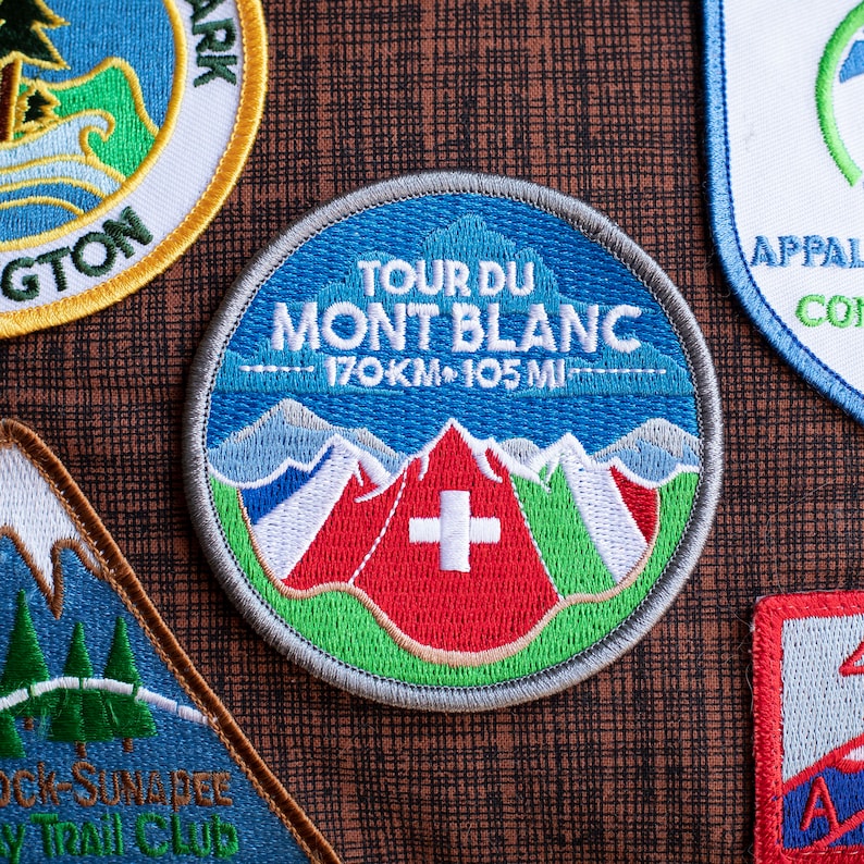 May include: Circular embroidered patch with the text "TOUR DU MONT BLANC" and "170KM 105 MI". The patch features a mountain range design with red, white, and green accents, and a white cross. The patch has a grey border.