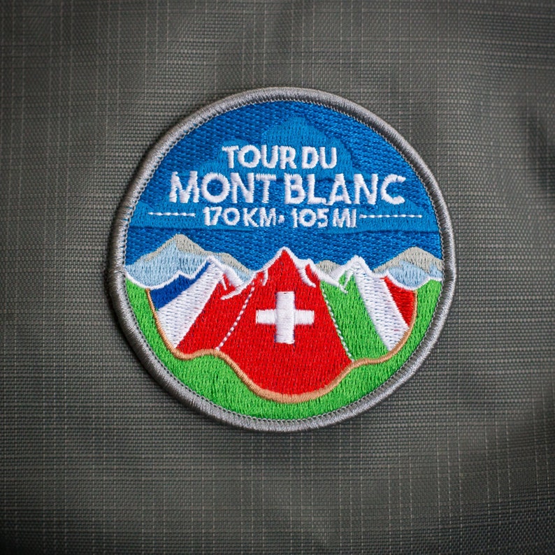 May include: Circular embroidered patch with the text "TOUR DU MONT BLANC" and the distance "170KM &bull; 105 MI". The design includes a blue sky, mountains, and a red mountain with a white cross, on a green and grey background.
