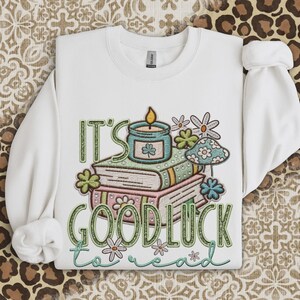 May include: White sweatshirt with the embroidered text "IT'S GOOD LUCK to read". The design includes a stack of books, a candle, flowers, and a mushroom. The design is in shades of green, pink, and blue.