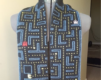 May include: A black knitted scarf with a blue and yellow Pac-Man maze design. The scarf features Pac-Man and ghosts in the maze.