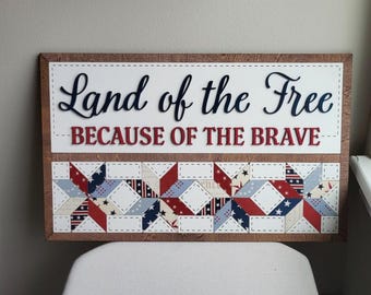 May include: Wooden sign with the text "Land of the Free BECAUSE OF THE BRAVE." The sign has a white background with black and red lettering. The bottom features a quilt-like design in red, white, and blue.