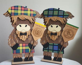 May include: Two wooden Highland cow figurines, each wearing a plaid tam and kilt. One holds a yellow banner reading "Clan Buchanan," the other a green banner reading "Clan MacDonald." The cows have brown fur, cream-colored faces, and black bow ties.