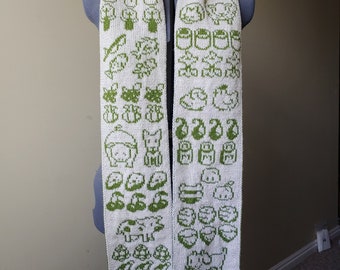 May include: A white knitted scarf with a green pixelated design of various animals and objects. The scarf is displayed on a white mannequin.