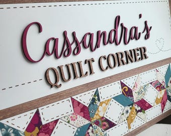 May include: A wooden sign with the text "Cassandra's QUILT CORNER" in a mix of cursive and block letters. The sign features a colorful quilt pattern at the bottom and a decorative border. The name is in a dark pink color.