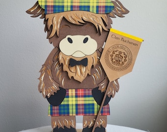 May include: A wooden Highland cow figurine wearing a plaid beret and kilt. The brown cow has a black bow tie and holds a banner that reads "Clan Buchanan". The figurine is on a wooden base.