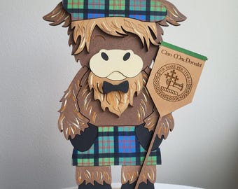 May include: A wooden Highland cow figure wearing a plaid hat and kilt. The cow has a brown body, a cream-colored face, and a black bow tie. It holds a banner that reads "Clan MacDonald" and features a crest. The figure stands on a wooden base.