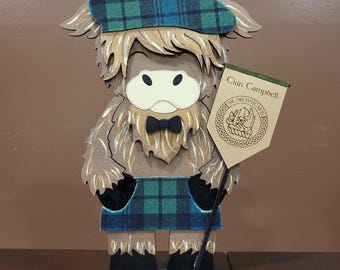 May include: A wooden Highland cow figurine wearing a plaid beret and kilt. The cow holds a banner that reads "Clan Campbell" with a circular emblem. The figurine has a black bow tie and is made of layered wood with a detailed design.