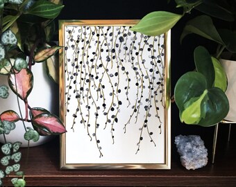 May include: A gold framed print of a black and white illustration of a cascading vine with small round berries. The print is displayed on a shelf with other plants.