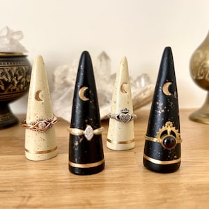 Könnte beinhalten: Vier dekorative Ringhalter. Zwei sind schwarz mit goldenen Akzenten, zwei cremefarben. Jeder hat ein Halbmond-Design und ein goldenes Band. Ringe verschiedener Designs werden auf den Haltern präsentiert.