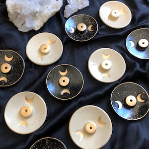 May include: A collection of ceramic incense holders in various colors, including black, white, and speckled designs. Each holder features a central hole and a gold crescent moon design. The holders are arranged on a dark, silky surface.