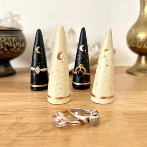 May include: Four decorative ring holders in black and cream colors, each with a gold crescent moon design. Two rings are displayed at the base of the holders. The holders are on a wooden surface.