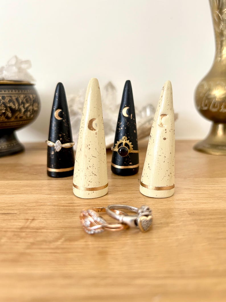 May include: Four decorative ring holders in black and cream colors, each with a gold crescent moon design. Two rings are displayed at the base of the holders. The holders are on a wooden surface.