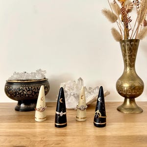 May include: Four ring holders with rings, each with a different design. Two are black with a gold base and a crescent moon. Two are cream-colored with a gold base and a crescent moon. A brass vase with dried flowers is in the background.