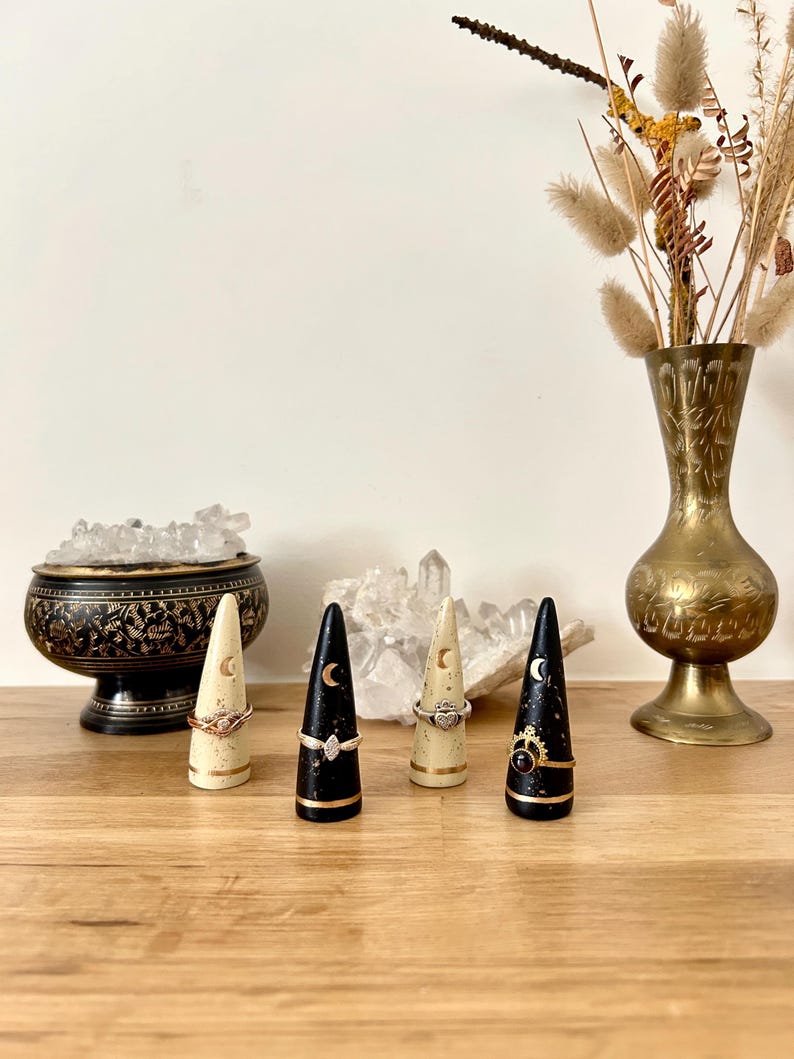 May include: Four ring holders with rings, each with a different design. Two are black with a gold base and a crescent moon. Two are cream-colored with a gold base and a crescent moon. A brass vase with dried flowers is in the background.