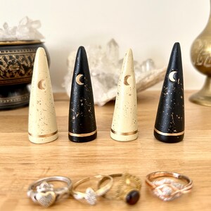 May include: Four wooden ring holders in cream and black with gold accents and crescent moon designs. Below are four rings with various designs. The ring holders are cone-shaped and sit on a wooden surface.