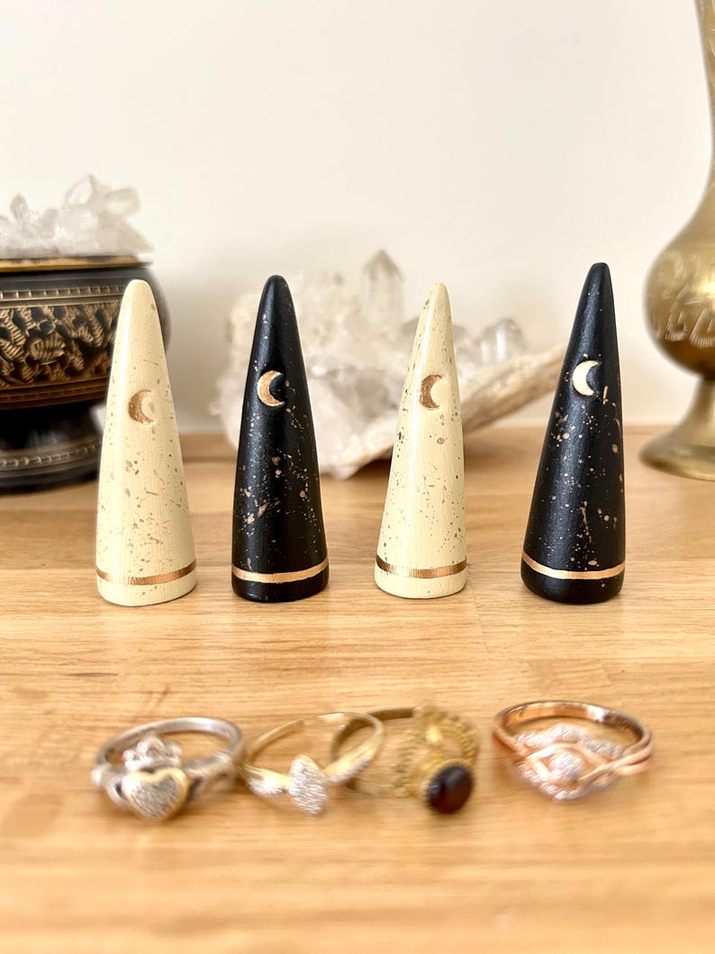May include: Four wooden ring holders in cream and black with gold accents and crescent moon designs. Below are four rings with various designs. The ring holders are cone-shaped and sit on a wooden surface.