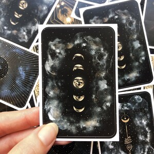 May include: A black card with a white border features a gold illustration of the moon in various phases against a black background with white clouds and gold glitter.
