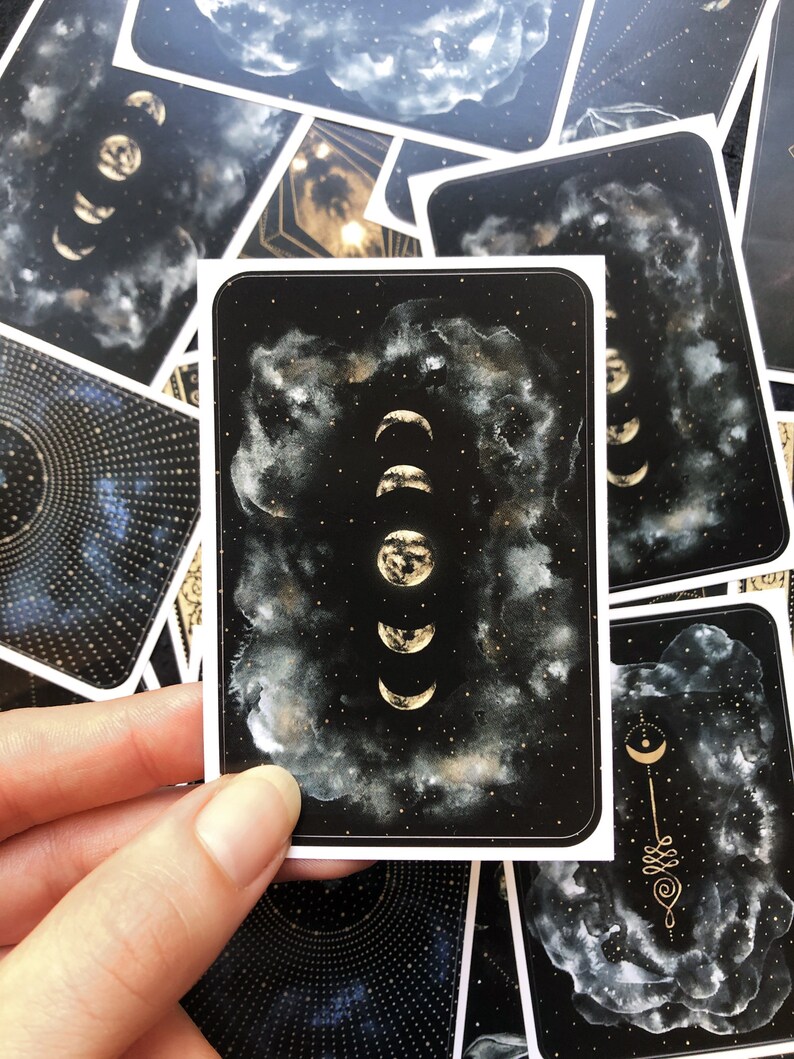 May include: A black card with a white border features a gold illustration of the moon in various phases against a black background with white clouds and gold glitter.