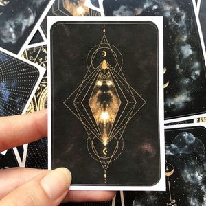 May include: A black card with a gold geometric design featuring a diamond shape with a crescent moon inside. The card has a dark background with a subtle starry pattern.