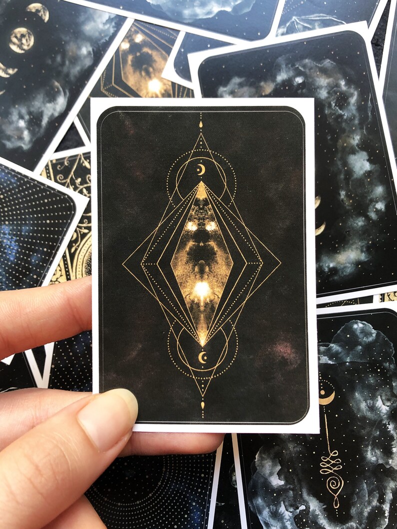 May include: A black card with a gold geometric design featuring a diamond shape with a crescent moon inside. The card has a dark background with a subtle starry pattern.