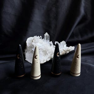May include: Four cone-shaped decorative objects, two black and two cream-colored, each with a crescent moon design and a copper-colored band. The objects are arranged in front of a cluster of clear quartz crystals against a black background.