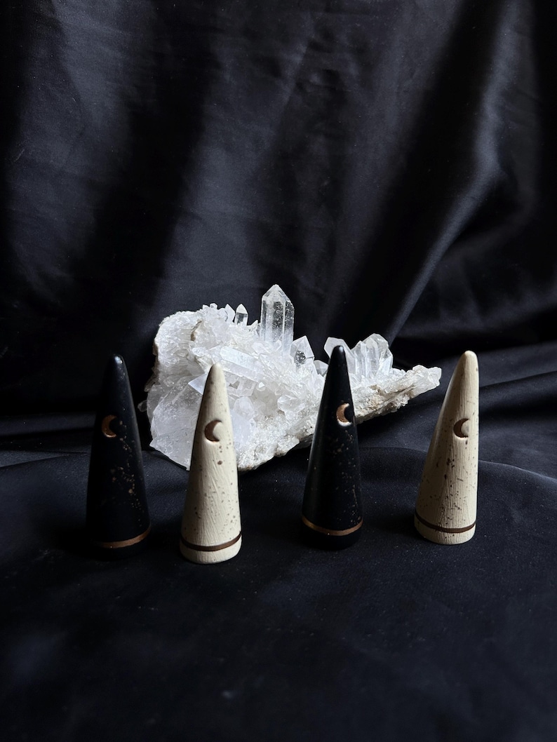 May include: Four cone-shaped decorative objects, two black and two cream-colored, each with a crescent moon design and a copper-colored band. The objects are arranged in front of a cluster of clear quartz crystals against a black background.